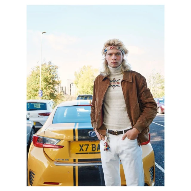 Man standing next to yellow Lexus car in parking lot. Man wears brown jacket, white jeans and headband with mulleted blond hair.