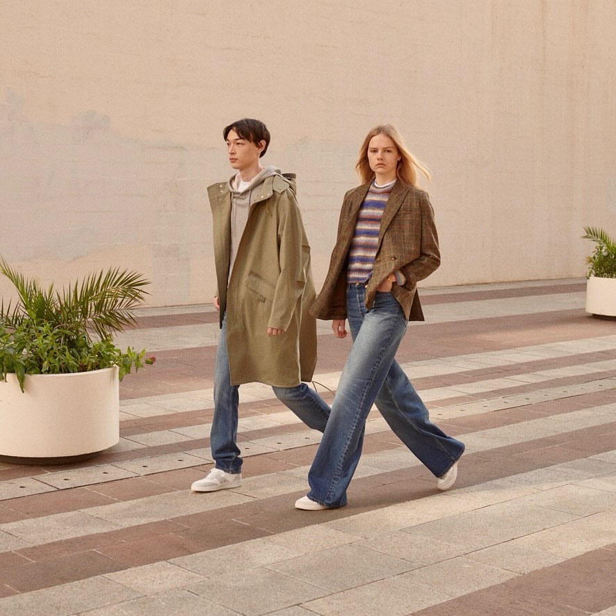 Two people walking in wide-leg jeans and jackets outdoors on a crosswalk in daylight. The man wears a parka and the woman a blazer.