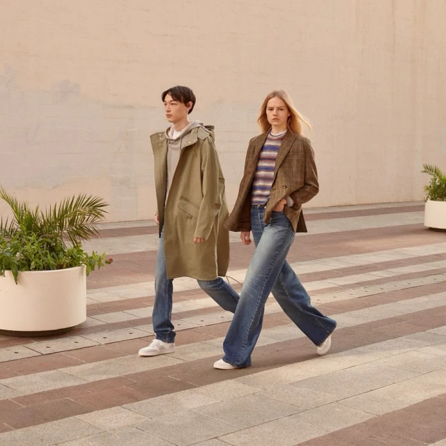 Two people walking in wide-leg jeans and jackets outdoors on a crosswalk in daylight. The man wears a parka and the woman a blazer.