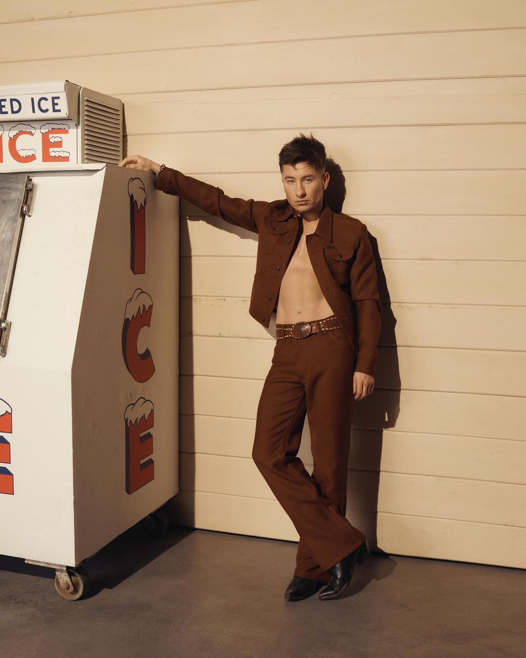 Portrait of a young adult man in a brown suit leaning against an ice machine with an open shirt, complemented by brown and neutral tones.