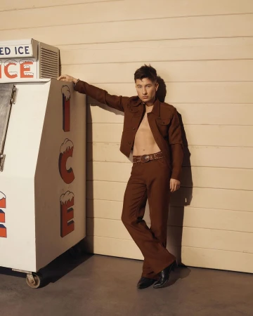 Portrait of a young adult man in a brown suit leaning against an ice machine with an open shirt, complemented by brown and neutral tones.