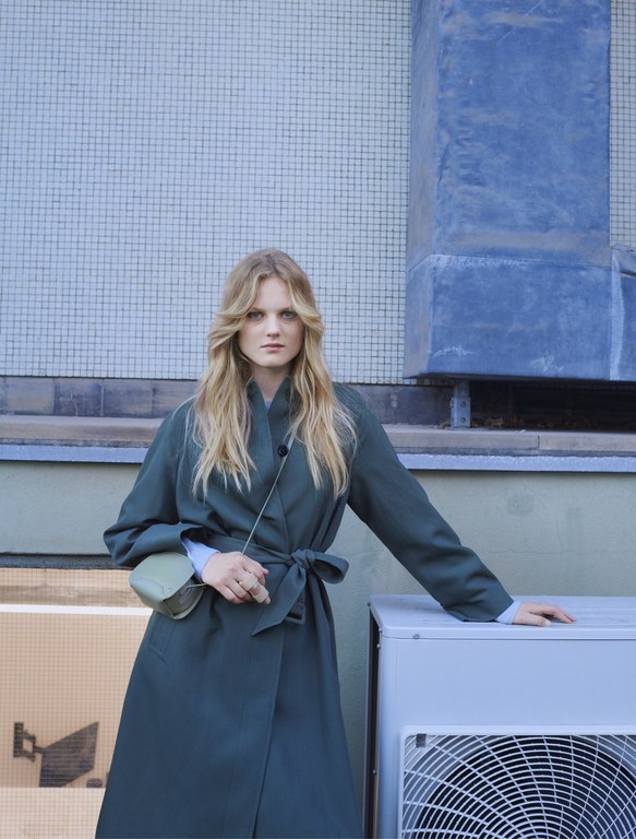 Adult female fashion model in a green trench coat poses outdoors against a tiled wall with a small purse and a neutral expression.