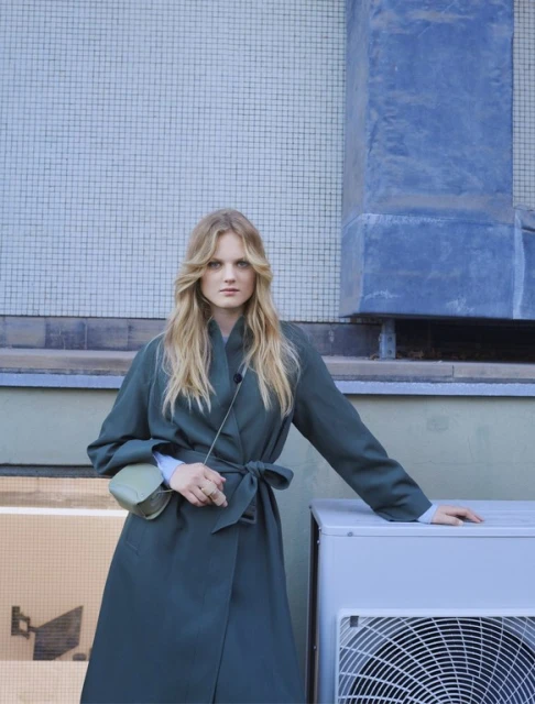 Adult female fashion model in a green trench coat poses outdoors against a tiled wall with a small purse and a neutral expression.