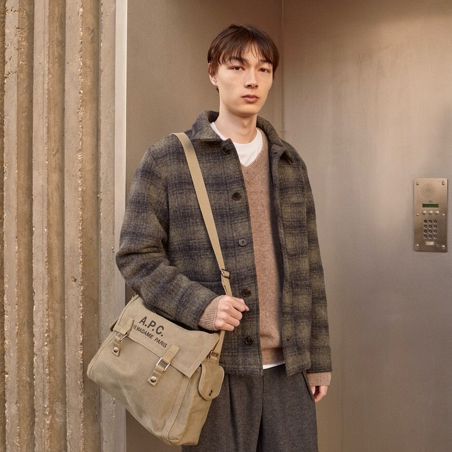 Male model wearing a plaid jacket, sweater, and grey pants carries a beige messenger bag in a neutral-toned indoor setting.