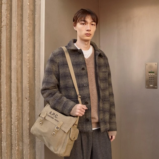 Male model wearing a plaid jacket, sweater, and grey pants carries a beige messenger bag in a neutral-toned indoor setting. 