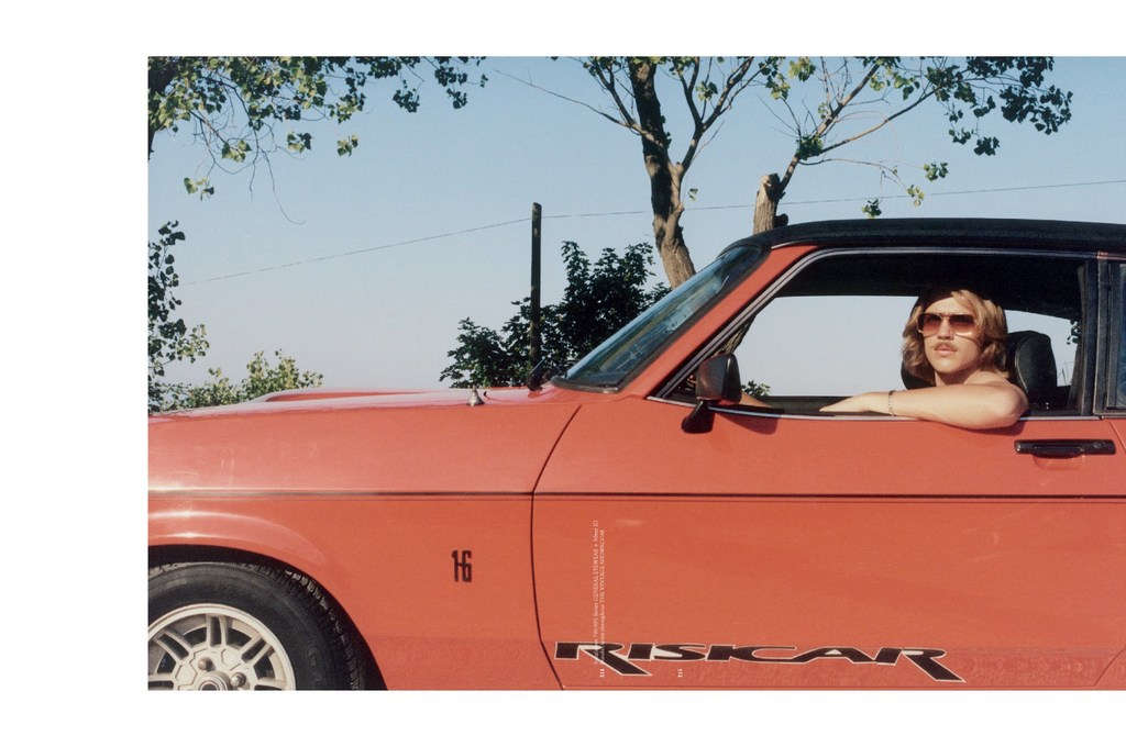Woman with sunglasses in a vintage red Opel Riskcar convertible with blue sky and sparse trees in the background on a sunny day.