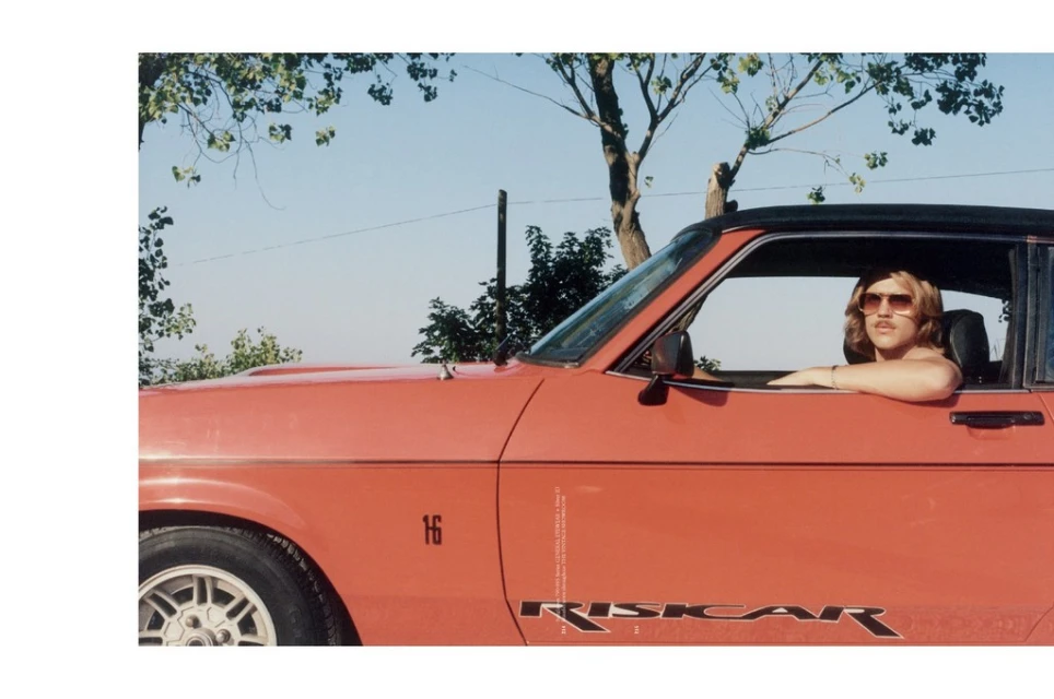 Woman with sunglasses in a vintage red Opel Riskcar convertible with blue sky and sparse trees in the background on a sunny day.