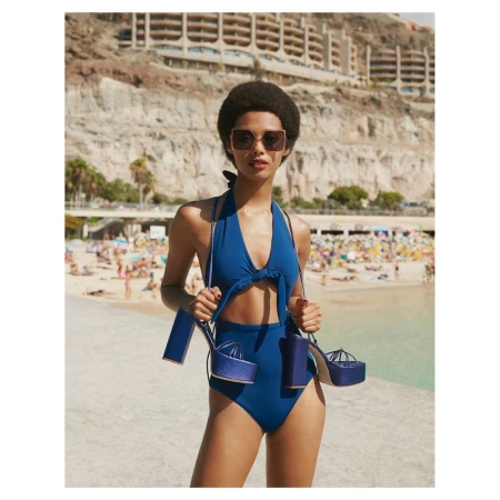 Model in blue swimsuit holding platform shoes outdoors near a beach with resort buildings in the background.