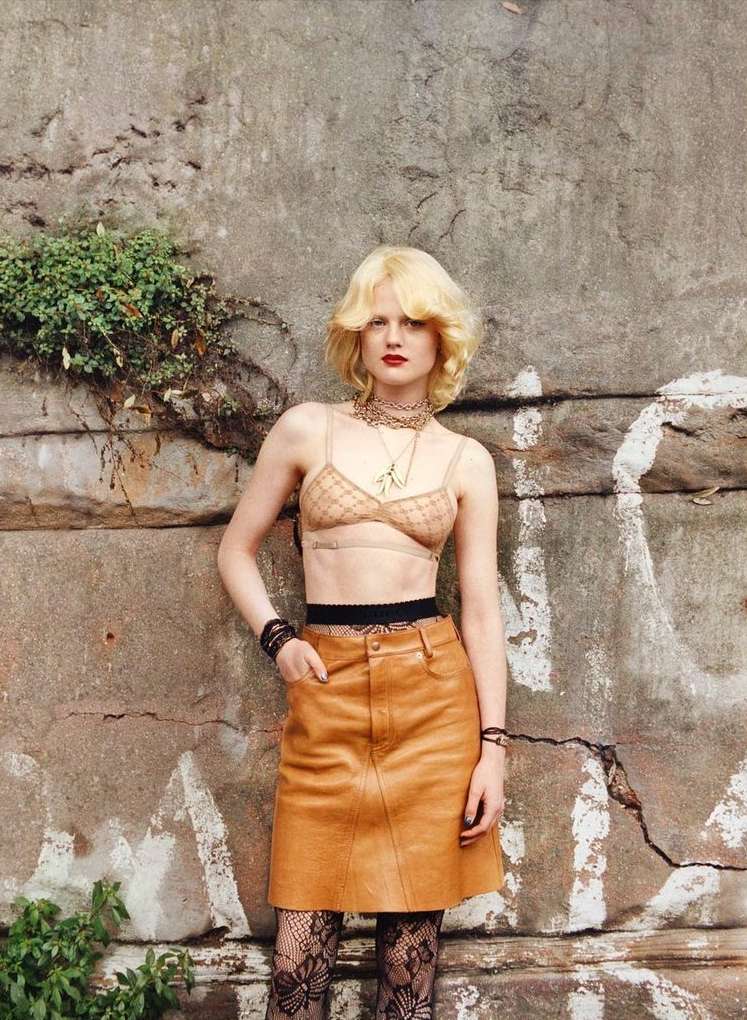 Young adult woman posing in a bra top, skirt, and patterned tights, standing in front of a weathered concrete wall with vegetation.