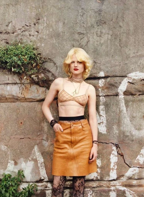 Young adult woman posing in a bra top, skirt, and patterned tights, standing in front of a weathered concrete wall with vegetation.