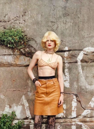 Young adult woman posing in a bra top, skirt, and patterned tights, standing in front of a weathered concrete wall with vegetation.