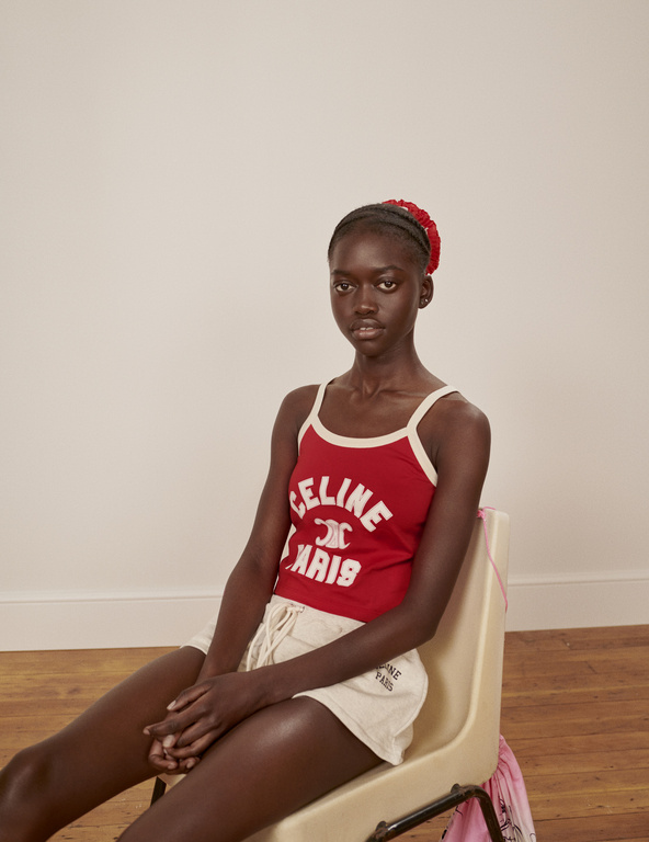 A young black woman with a red floral headband sits on a chair wearing a red tank top and shorts, posing in front of a neutral background.