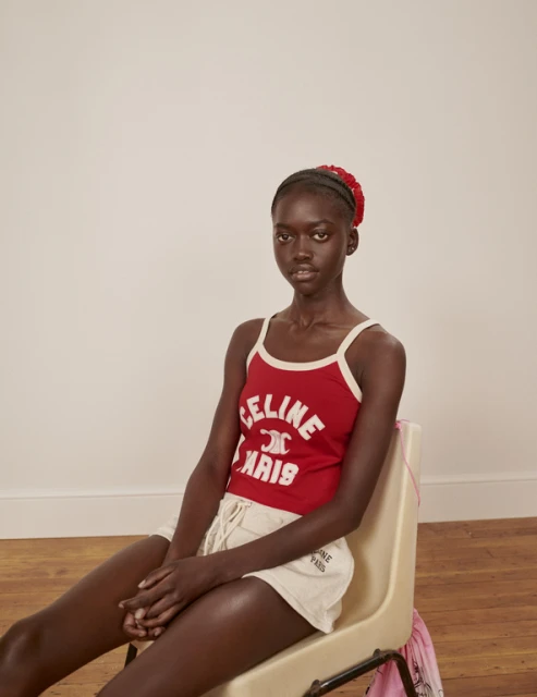 A young black woman with a red floral headband sits on a chair wearing a red tank top and shorts, posing in front of a neutral background.