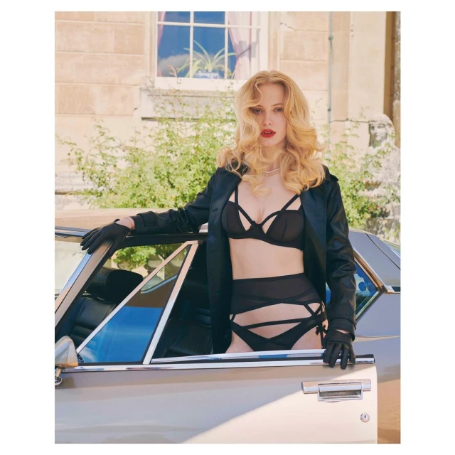 A woman with curly blonde hair, wearing lingerie and a black jacket, poses leaning against a vintage car outdoors on a sunny day.