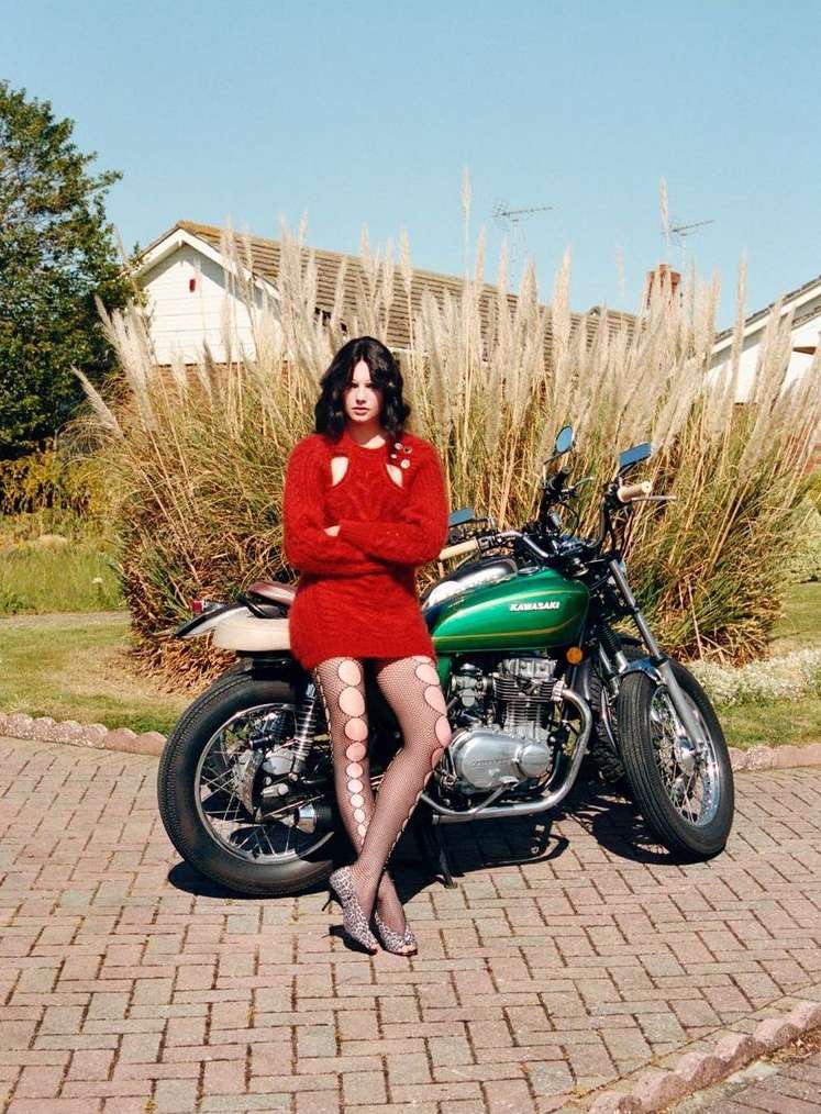 Brunette woman in a red knit dress poses next to a green Kawasaki motorcycle in a backyard setting on a sunny day.