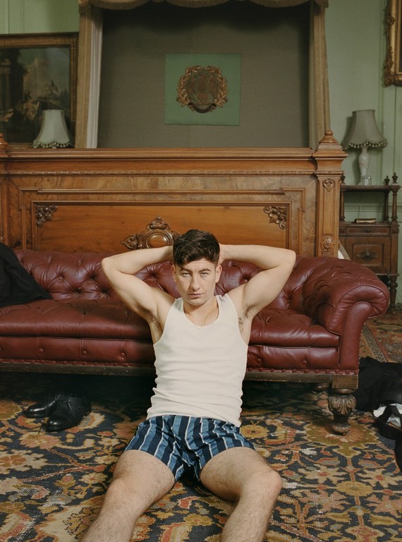 Young man with dark hair in tank top and blue striped shorts sits on patterned carpet with leather sofa and antique bed in the background.