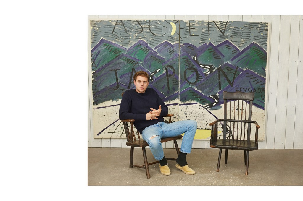 A young man seated in a wooden chair wearing a blue sweater and light wash jeans against a painted landscape backdrop styled by Venetia Scott