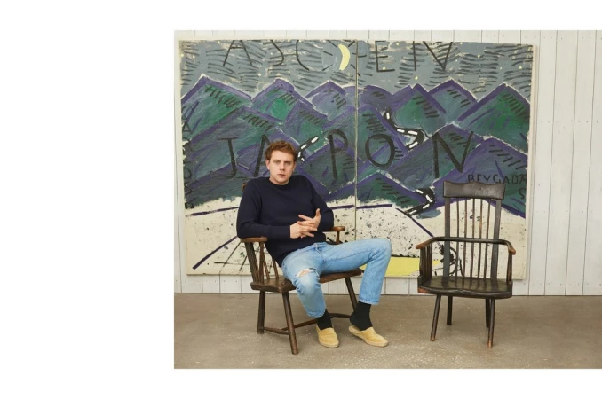A young man seated in a wooden chair wearing a blue sweater and light wash jeans against a painted landscape backdrop styled by Venetia Scott