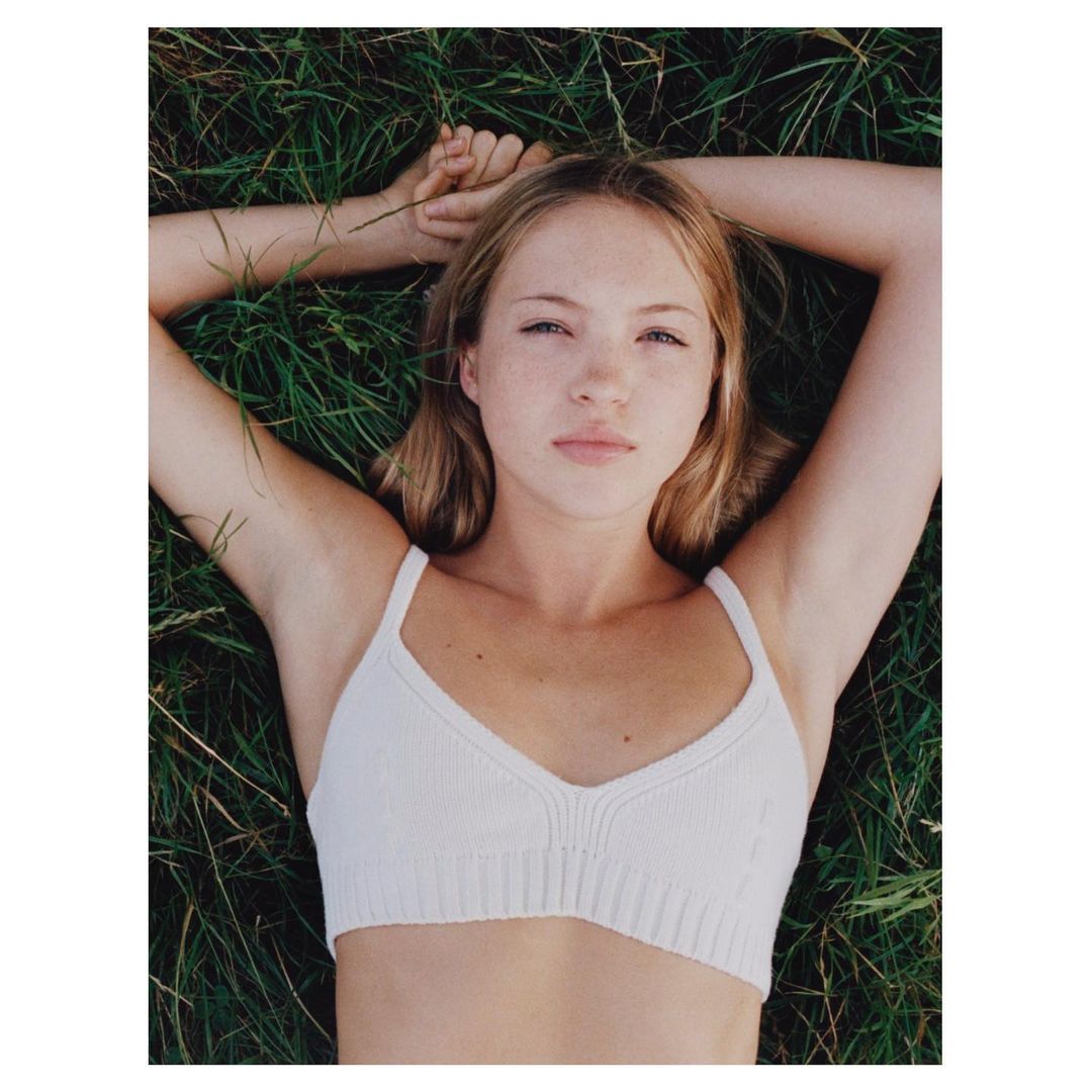 Young blonde woman lies on back in grass wearing a white knit top, looking up, natural lighting, beauty shot, and minimalist styling.
