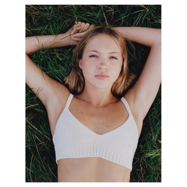 Young blonde woman lies on back in grass wearing a white knit top, looking up, natural lighting, beauty shot, and minimalist styling.