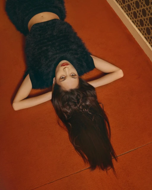 Overhead portrait of a woman in black fuzzy top and skirt laying on a orange carpet with dark hair spread across floor indoors.
