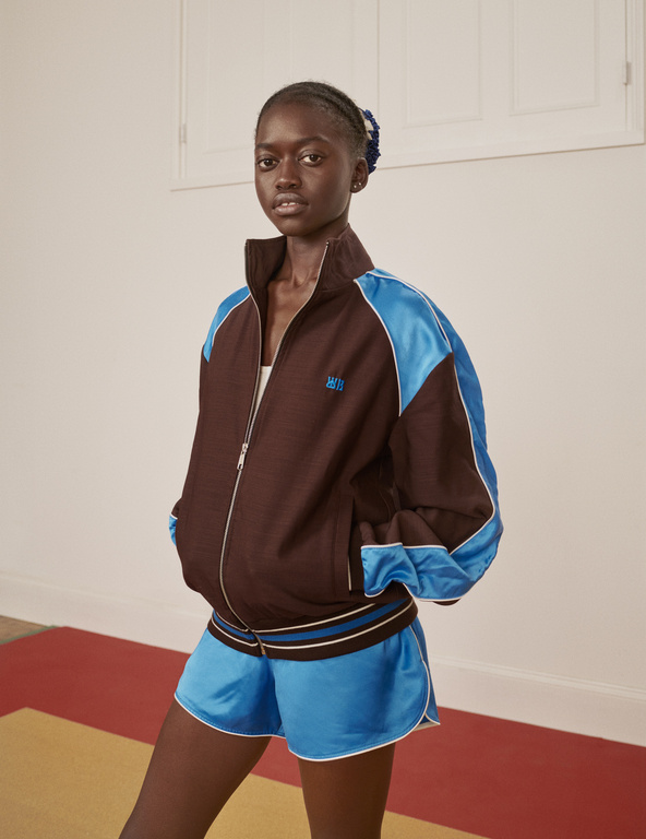 An African-American female model poses in a brown and blue retro-style track jacket and matching shorts in a neutral indoor setting.