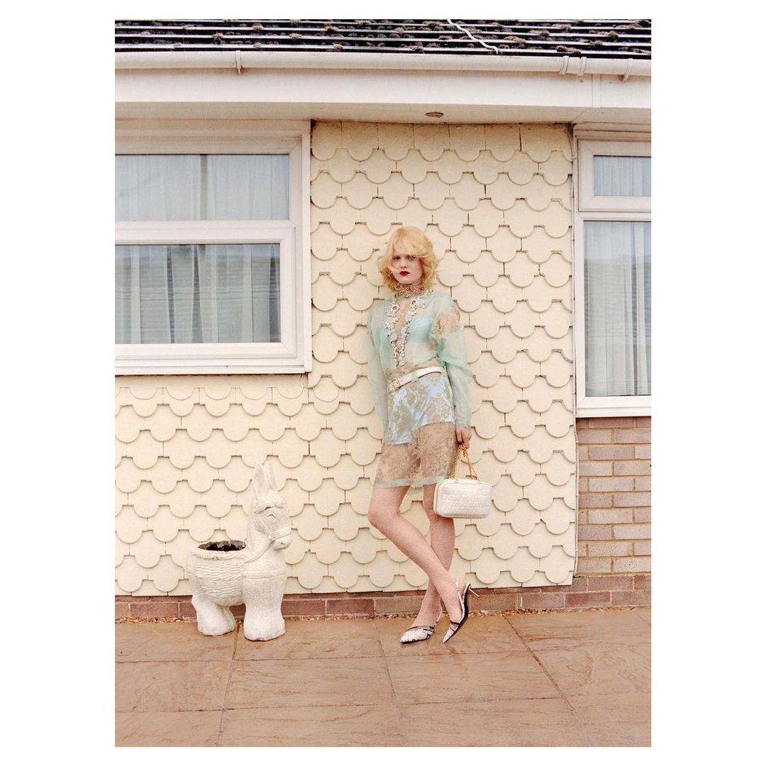 Woman in a sheer floral dress poses outside a house with an animal-shaped planter, holding a white purse and wearing two-tone pumps.