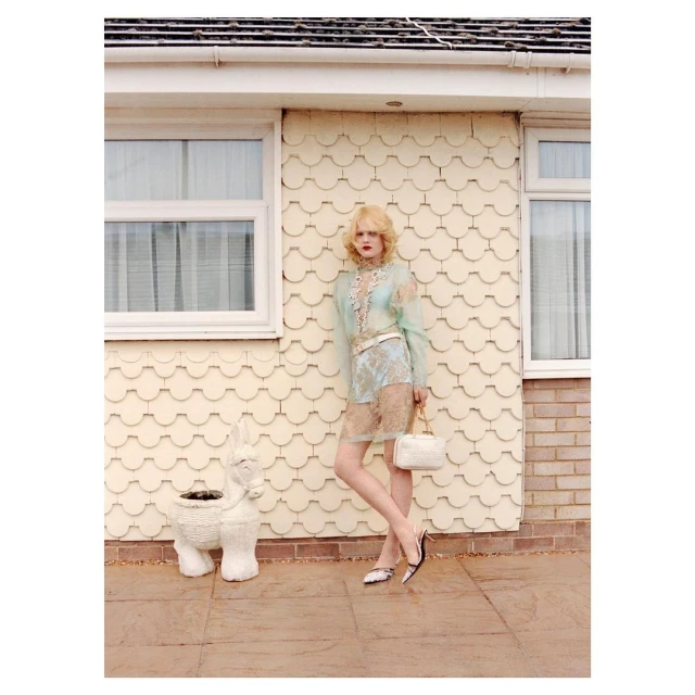 Woman in a sheer floral dress poses outside a house with an animal-shaped planter, holding a white purse and wearing two-tone pumps.