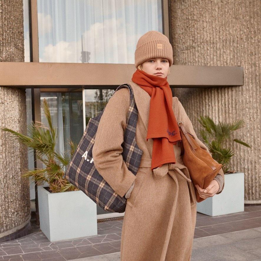 Woman in tan wool coat, beanie, and orange scarf carries tote bag near building exterior; fashion editorial with neutral tones and natural light.