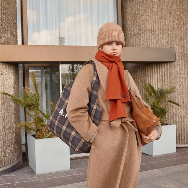 Woman in tan wool coat, beanie, and orange scarf carries tote bag near building exterior; fashion editorial with neutral tones and natural light.