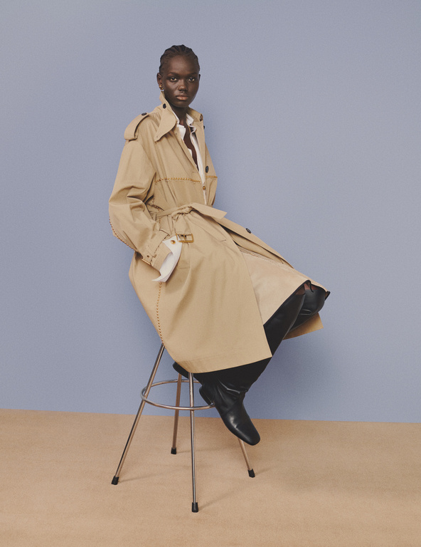 A fashion portrait of an adult female model in a trench coat, seated on a metal stool against a blue and beige background in a studio.