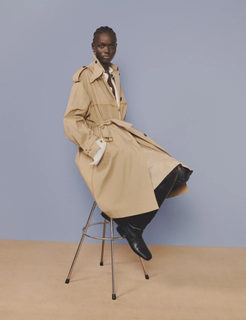 A fashion portrait of an adult female model in a trench coat, seated on a metal stool against a blue and beige background in a studio.