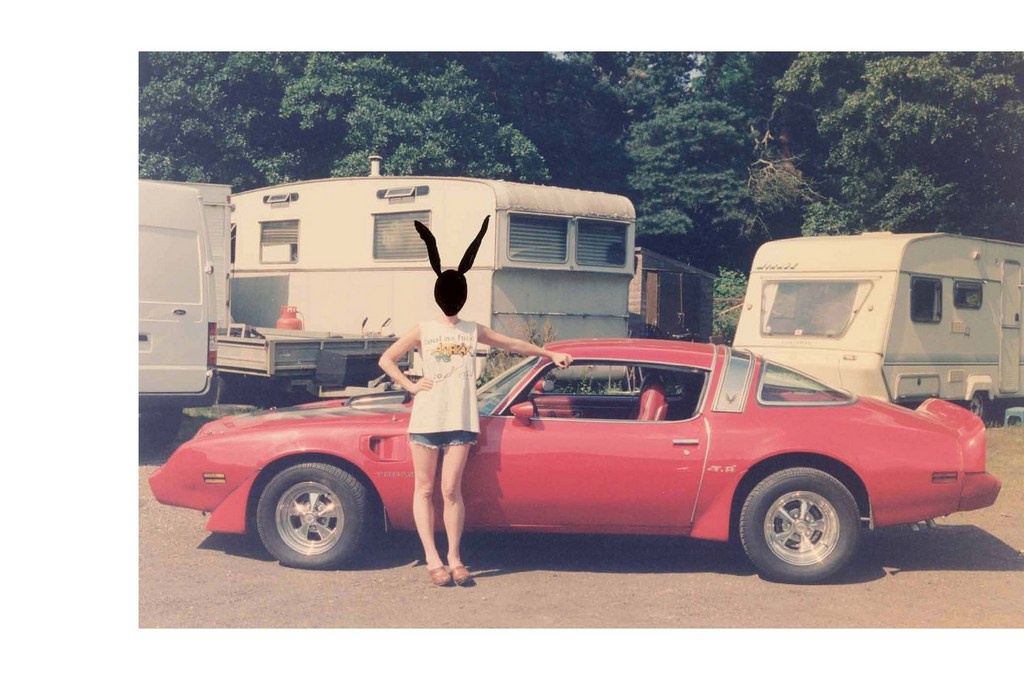 Person with rabbit ears standing by red sports car in front of vintage caravans on grassy lot under sunny skies.