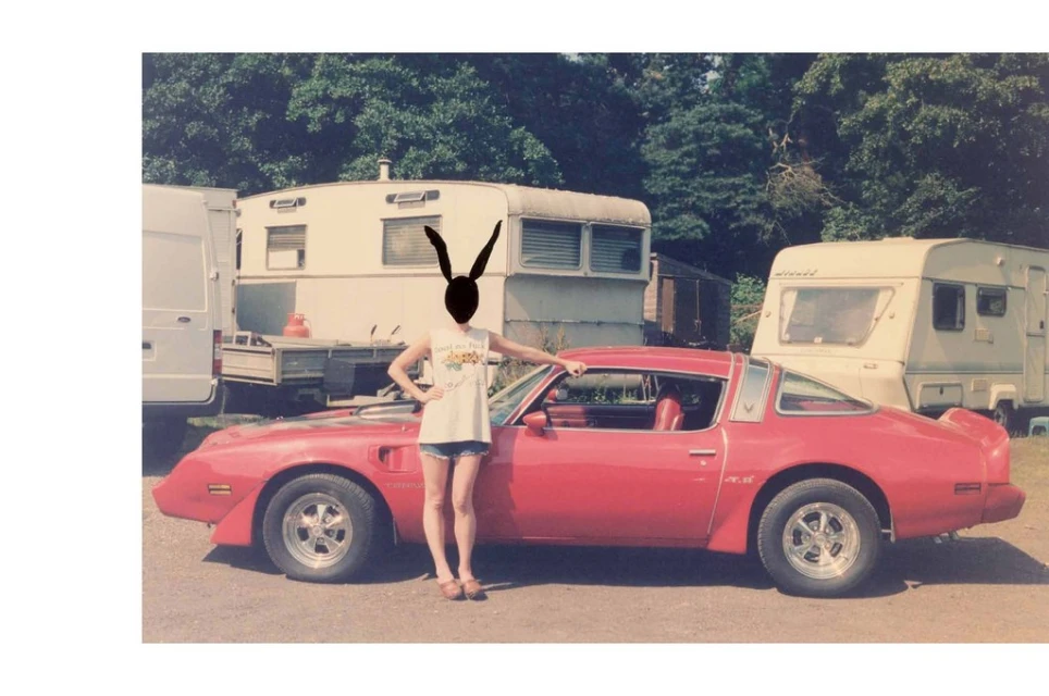 Person with rabbit ears standing by red sports car in front of vintage caravans on grassy lot under sunny skies.