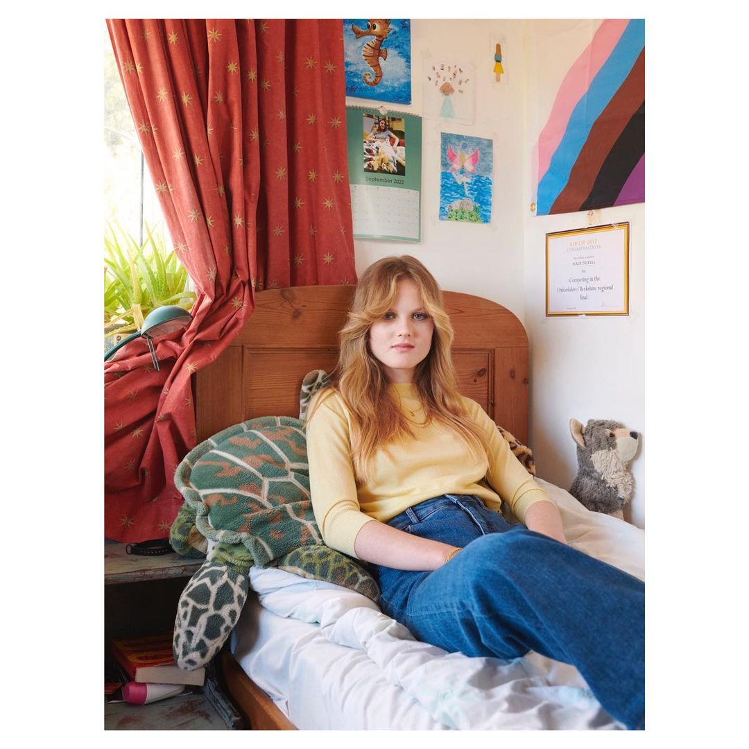 A woman with long blond hair sits on a bed next to a stuffed turtle in a bedroom with colorful artwork and red curtains in natural light.
