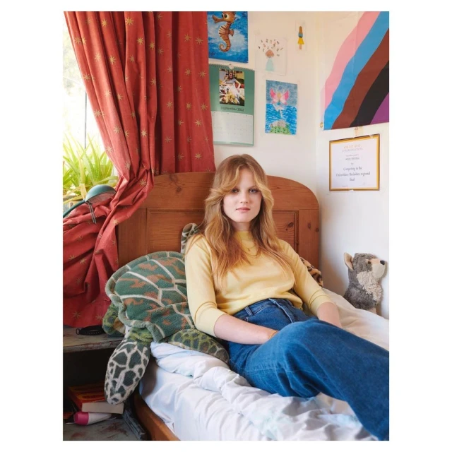 A woman with long blond hair sits on a bed next to a stuffed turtle in a bedroom with colorful artwork and red curtains in natural light.