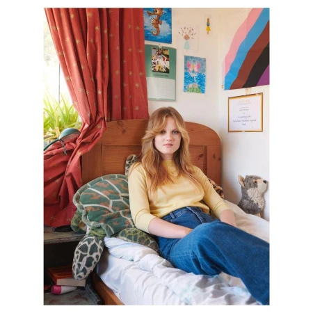 A woman with long blond hair sits on a bed next to a stuffed turtle in a bedroom with colorful artwork and red curtains in natural light.