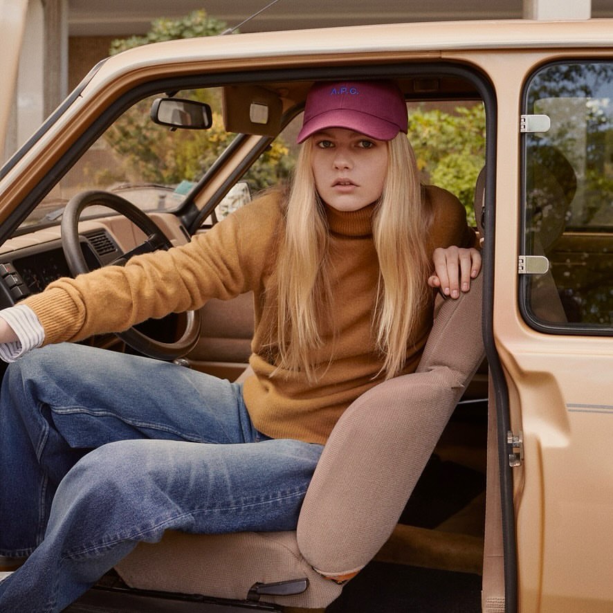 A blonde woman wearing a maroon cap and a tan sweater poses in the driver's seat of a car. The background features a house and trees.
