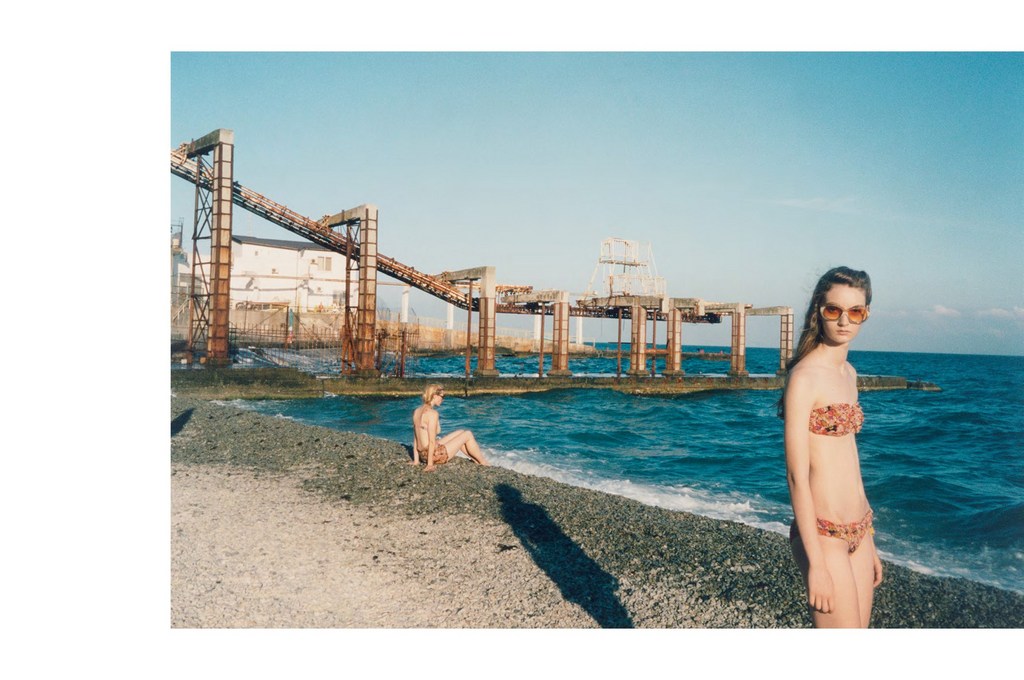 Two women in swimwear pose on a rocky beach with turquoise water and a dilapidated industrial structure in the background on a sunny day.
