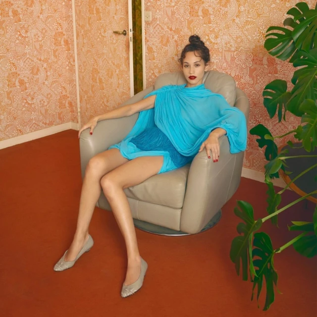 Adult woman in a blue dress sitting in a chair with a monstera plant, interior setting, and wallpapered wall in the background.
