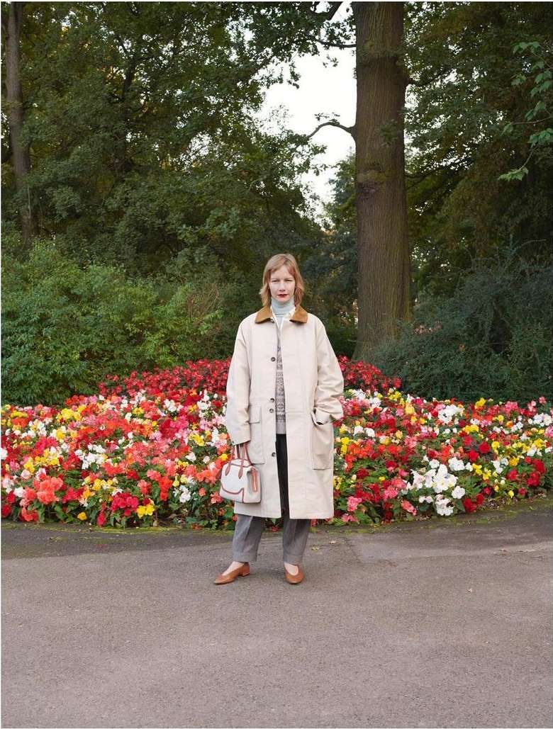 A woman stands in front of a colorful flowerbed in a park-like setting, wearing a beige coat and carrying a small handbag.