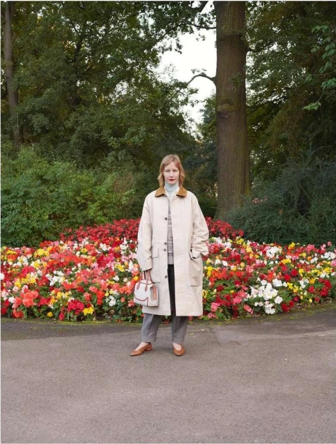 A woman stands in front of a colorful flowerbed in a park-like setting, wearing a beige coat and carrying a small handbag.