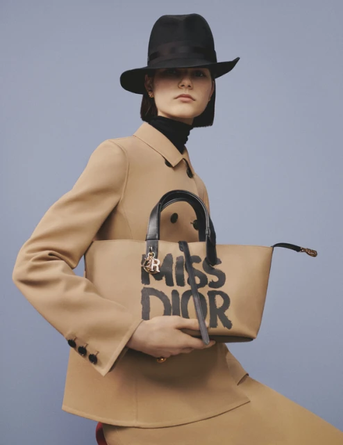 Elegant woman in a tan skirt suit and black hat holding a Miss Dior handbag against a light blue background in a fashion editorial.