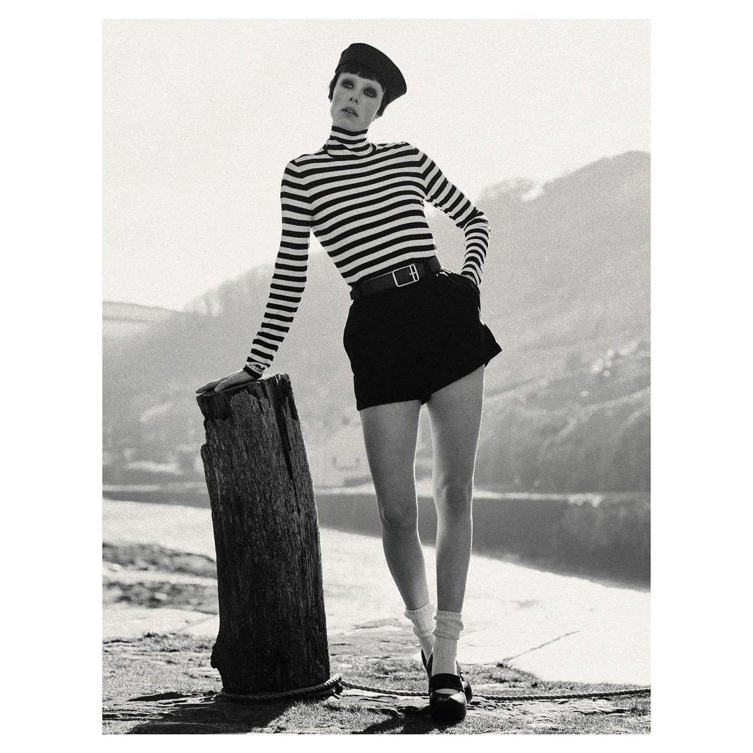 Monochrome portrait of woman in striped top, black shorts, and Mary Jane shoes, leaning against a wooden post with water in background.