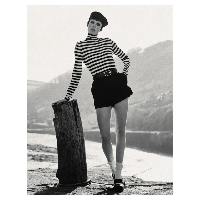 Monochrome portrait of woman in striped top, black shorts, and Mary Jane shoes, leaning against a wooden post with water in background.