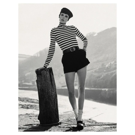 Monochrome portrait of woman in striped top, black shorts, and Mary Jane shoes, leaning against a wooden post with water in background.