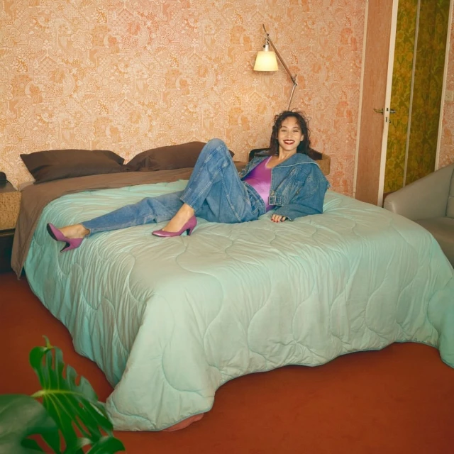 A young woman lounges on a bed in a vintage-style room with patterned wallpaper, wearing a denim jacket, jeans, and pink heels.