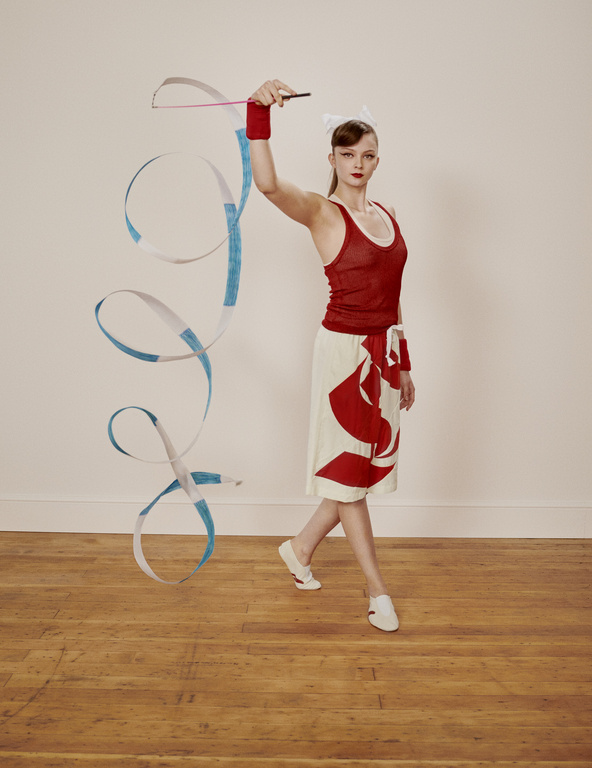 A woman performs rhythmic gymnastics with a ribbon, wearing a red top and patterned skirt, on a wooden floor against a plain background.