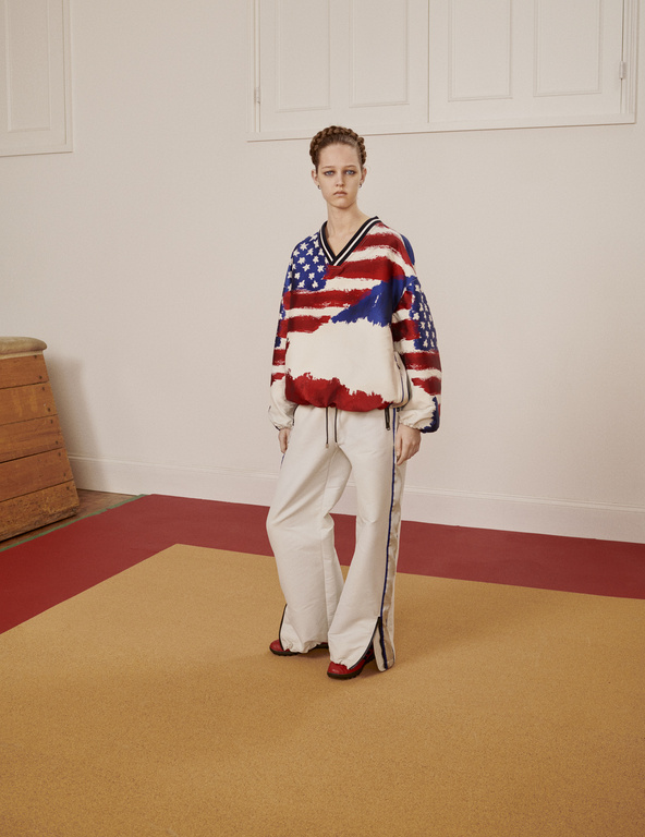 A woman in a patriotic-themed sweatshirt and track pants stands in a photo studio. The walls are painted white with a tan and red floor.