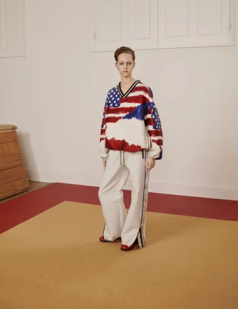 A woman in a patriotic-themed sweatshirt and track pants stands in a photo studio. The walls are painted white with a tan and red floor.